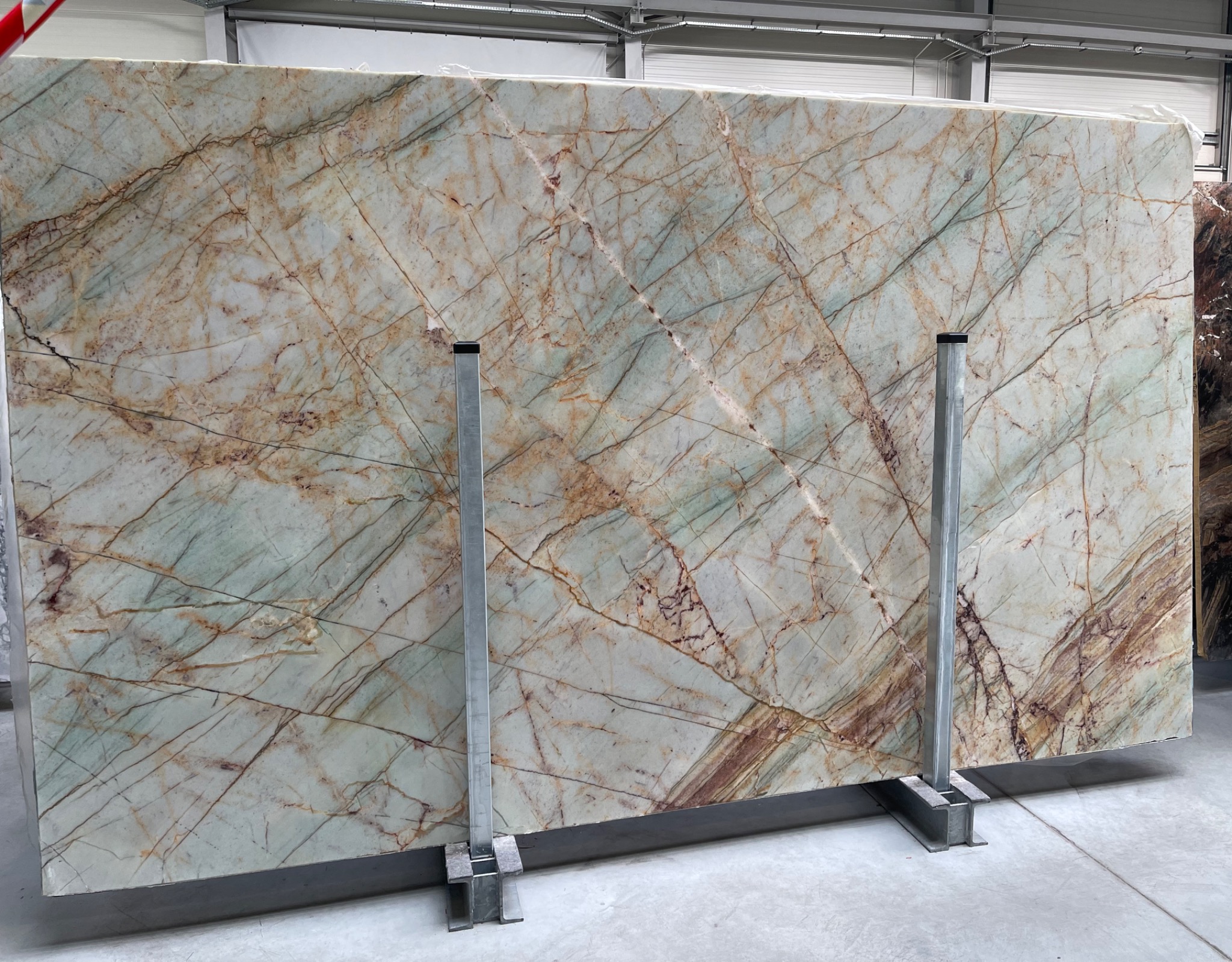 Duża płyta kwarcu brazylijskiego z wyraźnym wzorem żył w odcieniach brązu i zieleni, podparta metalowymi wspornikami w magazynie.