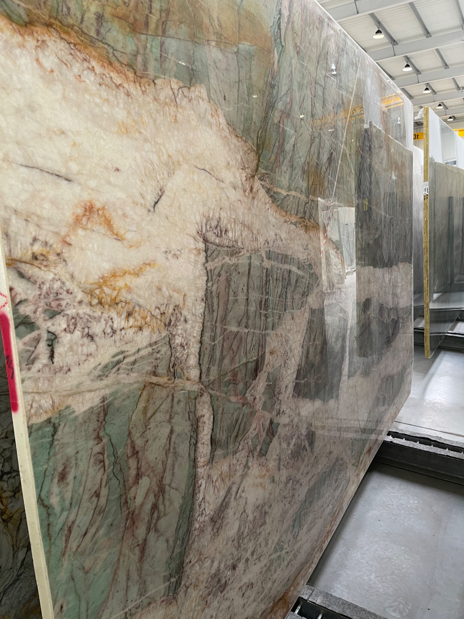Wielkoformatowa płyta z kamienia naturalnego o unikalnym wzorze z przewagą bieli, zieleni i brązu, ustawiona pionowo w magazynie.