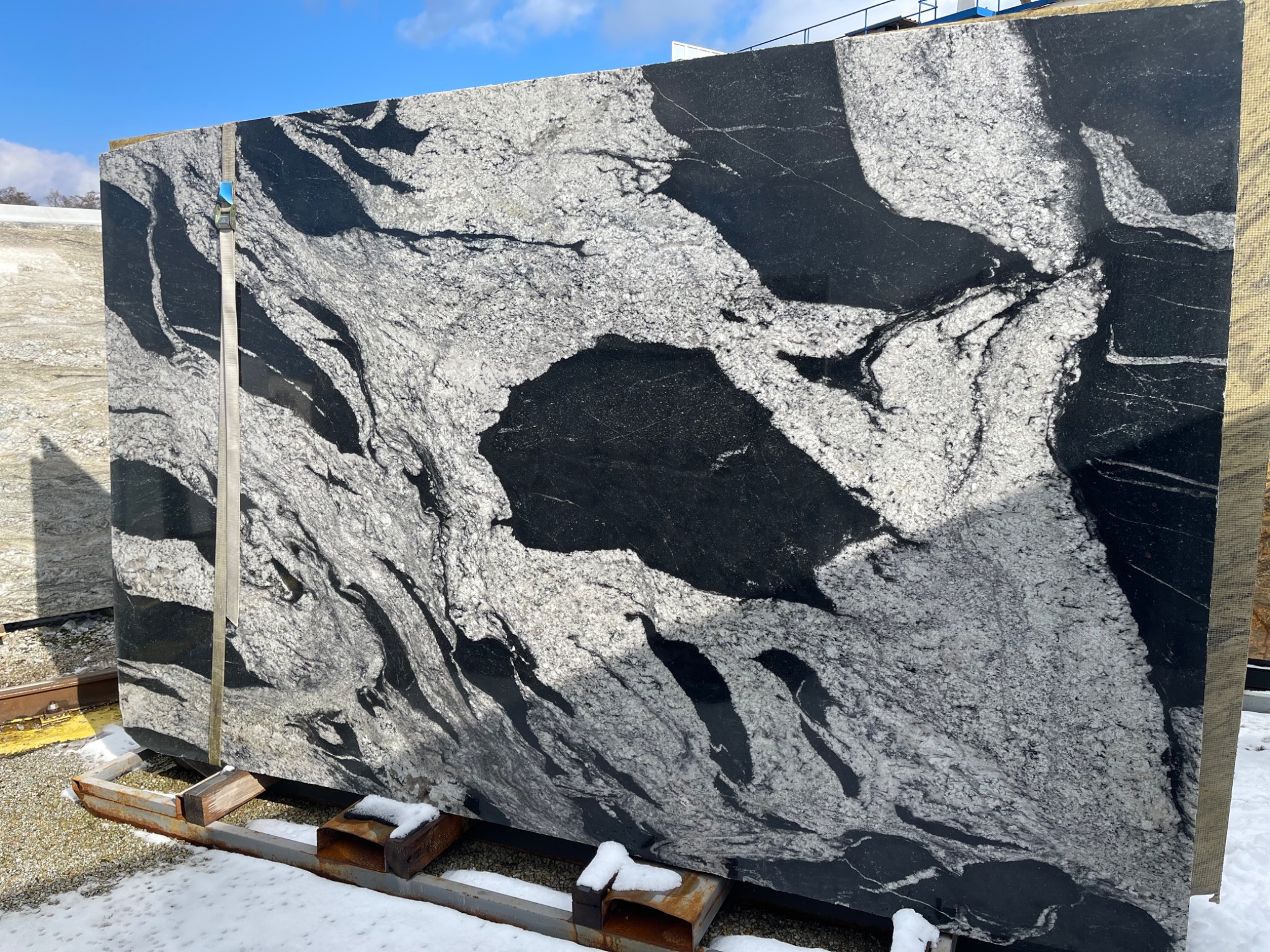Duża płyta kamienna z czarno-białym wzorem żyłkowania, zabezpieczona pasem transportowym, oparta na metalowej ramie na zewnątrz w zimowych warunkach.