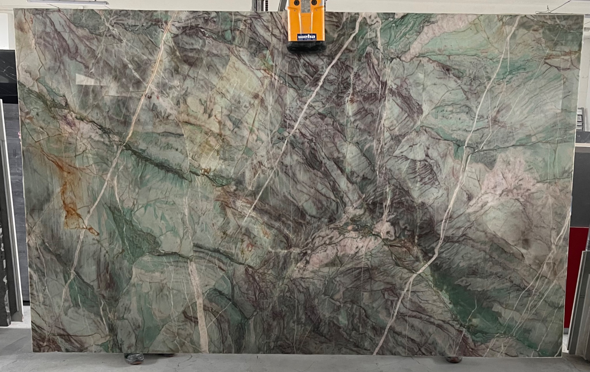 Duża płyta kamienna o zielono-szarym marmurkowym wzorze, zabezpieczona żółtym uchwytem próżniowym na górze.