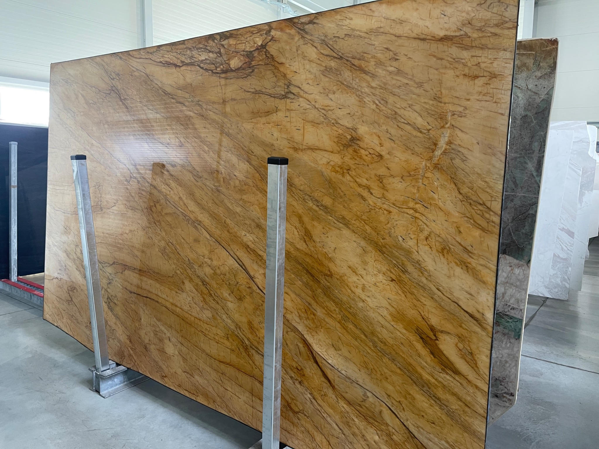 Duża płyta kamienna w odcieniach brązu i beżu z wyraźnym, naturalnym wzorem żyłkowania, oparta o metalowe wsporniki w magazynie, widoczne kolejne płyty w tle.