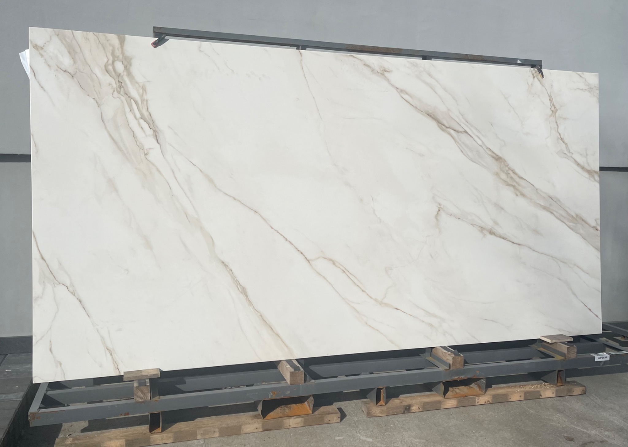 Duża płyta z białego kamienia z delikatnymi, brązowymi żyłami, oparta na metalowej konstrukcji z drewnianymi podkładkami, prezentowana w magazynie.