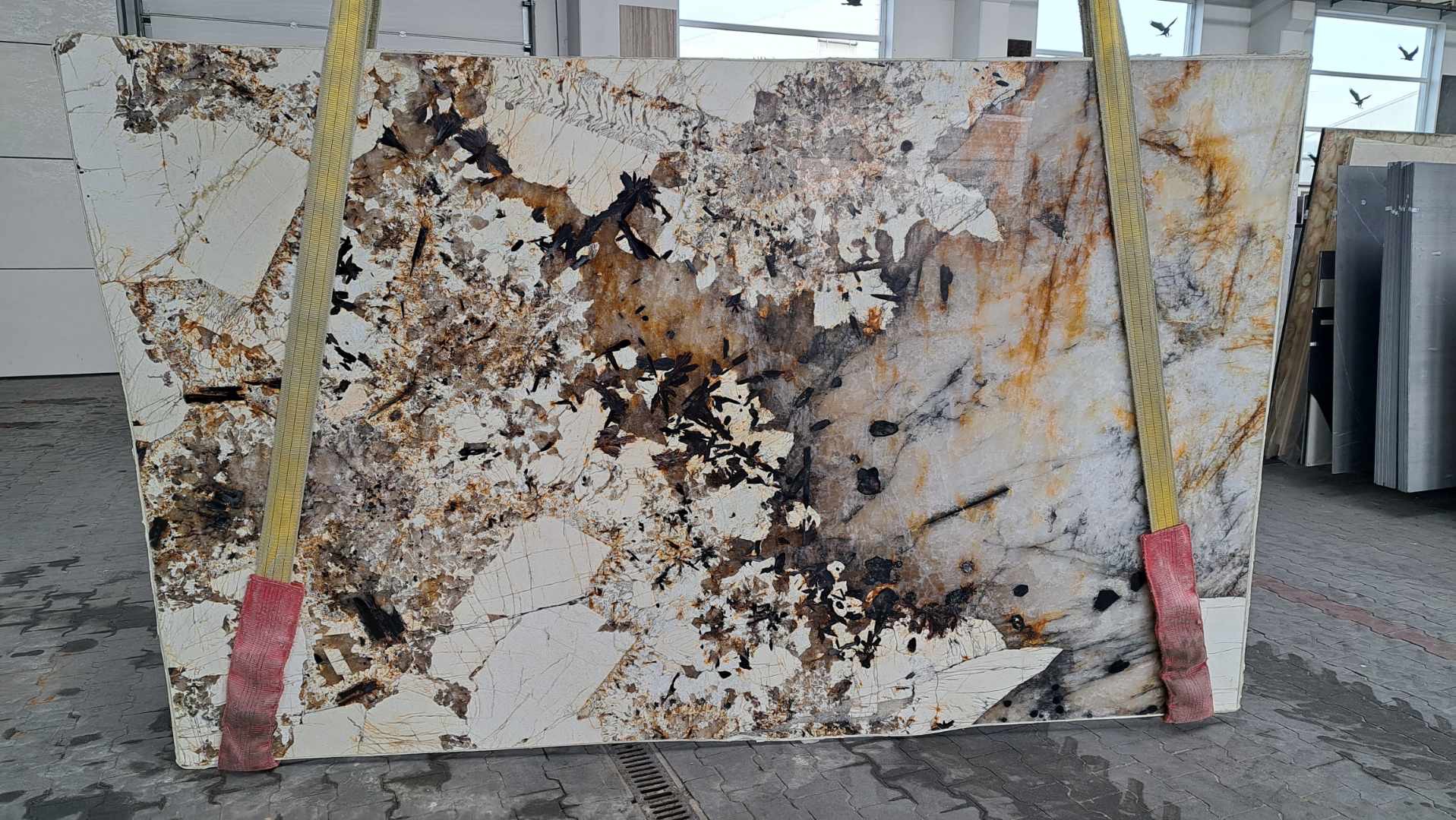 Płyta kwarcu brazylijskiego o nieregularnym wzorze, podwieszona na pasach transportowych w magazynie hurtowni kamienia.