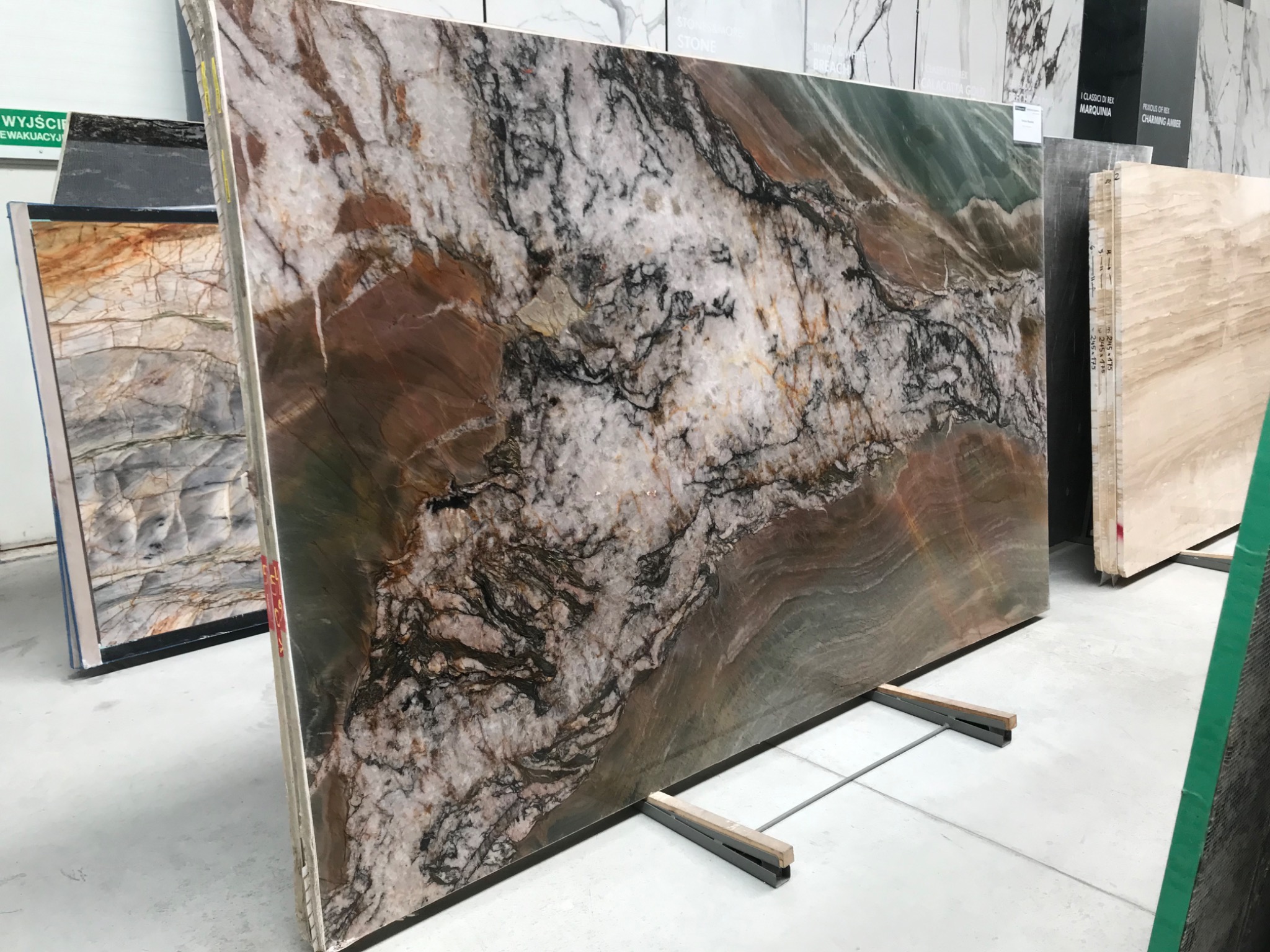 Duża płyta kamienna z unikalnym wzorem w odcieniach bieli, brązu, czerni i zieleni, ustawiona pionowo na metalowych wspornikach w magazynie kamieni.