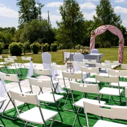 Ustawienie krzeseł i dekoracji na zewnątrz, przygotowane na ceremonię ślubną z różowym łukiem i stołem w tle.