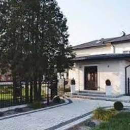 Nowoczesny dom jednorodzinny z jasną elewacją, ciemnym dachem i zadbanym ogrodem z podjazdem wyłożonym kostką brukową.