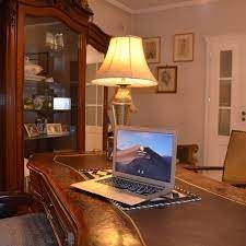 Biurko z ciemnego drewna w klasycznym stylu, na nim otwarty laptop z widokiem górskim na ekranie, w tle przeszklona witryna z dekoracjami i lampa z abażurem.