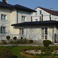 Jasny, dwupiętrowy dom z ciemnym dachem i białymi balustradami, otoczony zadbanym ogrodem z niskimi krzewami i ścieżką wyłożoną kamieniami.