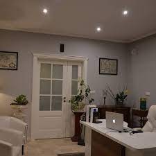 Wnętrze biura z białymi meblami i laptopem na biurku, oświetlone punktowymi lampami sufitowymi, z dekoracjami roślinnymi i obrazami na ścianach.