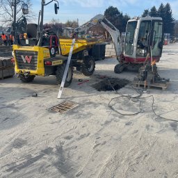 Rentbud Damian Kołodziej - Żółty wozidło i mini koparka Takeuchi TB216 na placu budowy z kostką brukową, widoczny wykop z kablami i studzienka kanalizacyjna, poziomica oparta o koło wozidła.