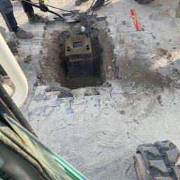 Rentbud Damian Kołodziej - Ekipa budowlana kopie dół pod betonowy element na placu wyłożonym szarą kostką, widoczny fragment traktora i poziomica.