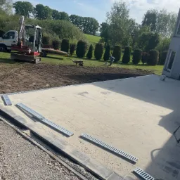 Przygotowany teren pod wylewkę z widocznym drenażem liniowym; w tle minikoparka i uporządkowany teren zielony. Jasne, słoneczne oświetlenie.