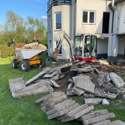 Rentbud Damian Kołodziej - Mini koparka i wywrotka na trawniku obok budynku w trakcie prac ziemnych. Stos gruzu i płyt betonowych. Widok na dom z oknami i drzewa w tle. Słoneczny dzień.