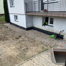 Rentbud Damian Kołodziej - Czarna folia izolacyjna przy fundamencie domu, obsypana żwirem. Obok stoi zagęszczarka. Widoczny fragment chodnika z kostki i łyżka koparki.