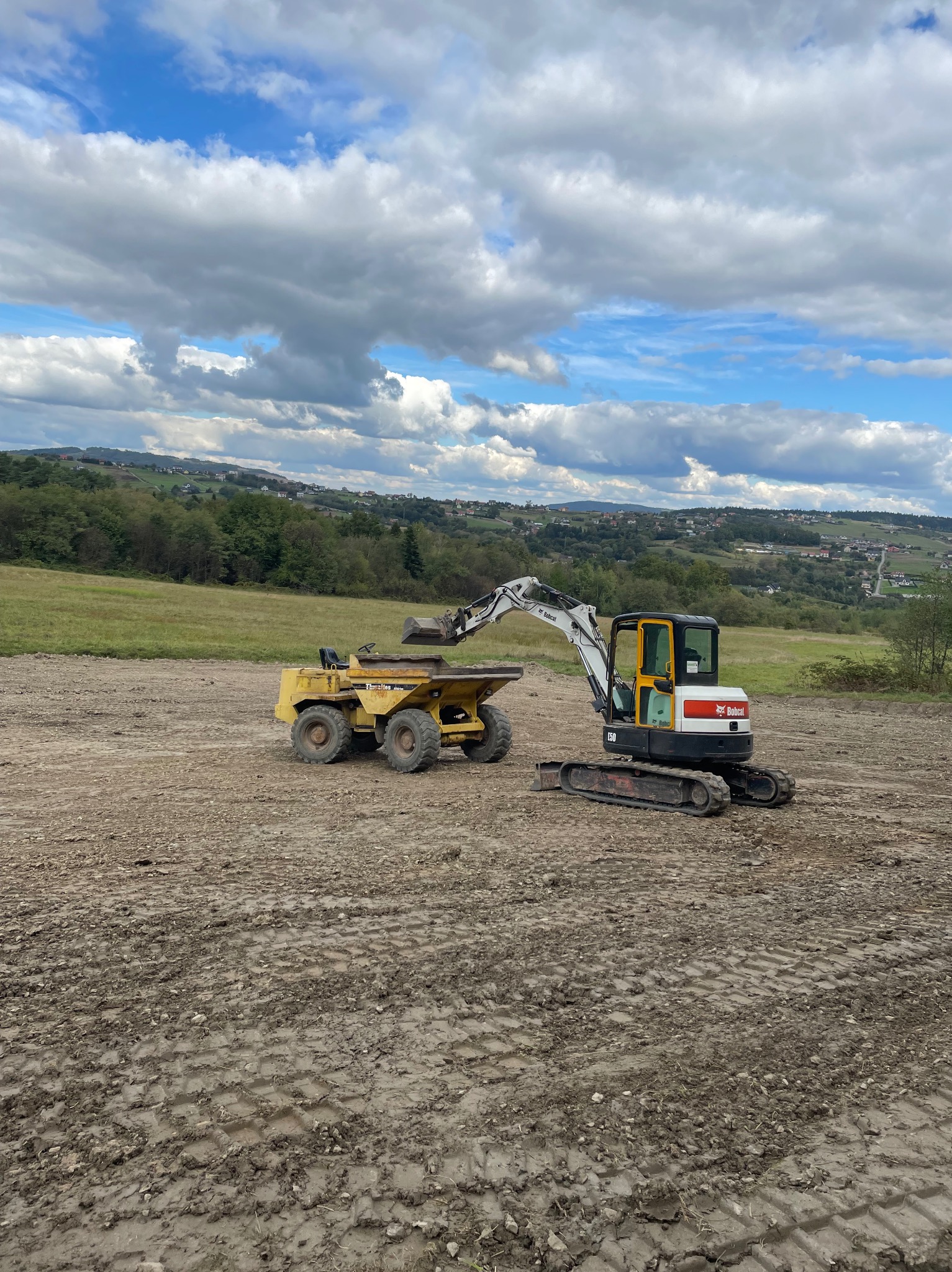 Żółty wywrotka i biało-czarny minikoparka Bobcat na placu budowy z widokiem na pagórkowaty krajobraz i zachmurzone niebo.