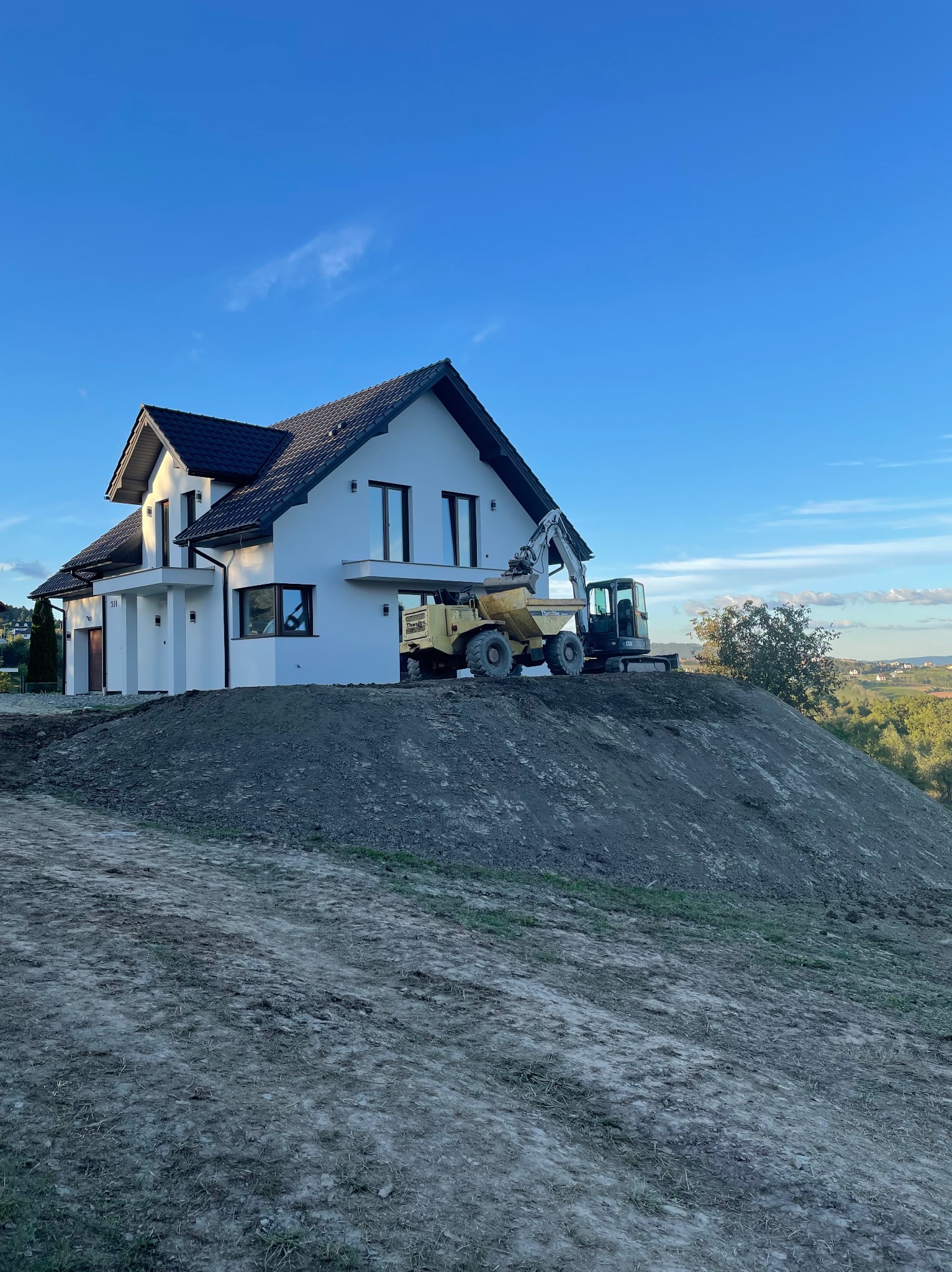 Nowoczesny dom jednorodzinny z wyeksponowanym nasypem ziemi, na którym stoi żółta wywrotka i mała koparka, sugerujące prace przygotowawcze pod ogród lub teren zielony.