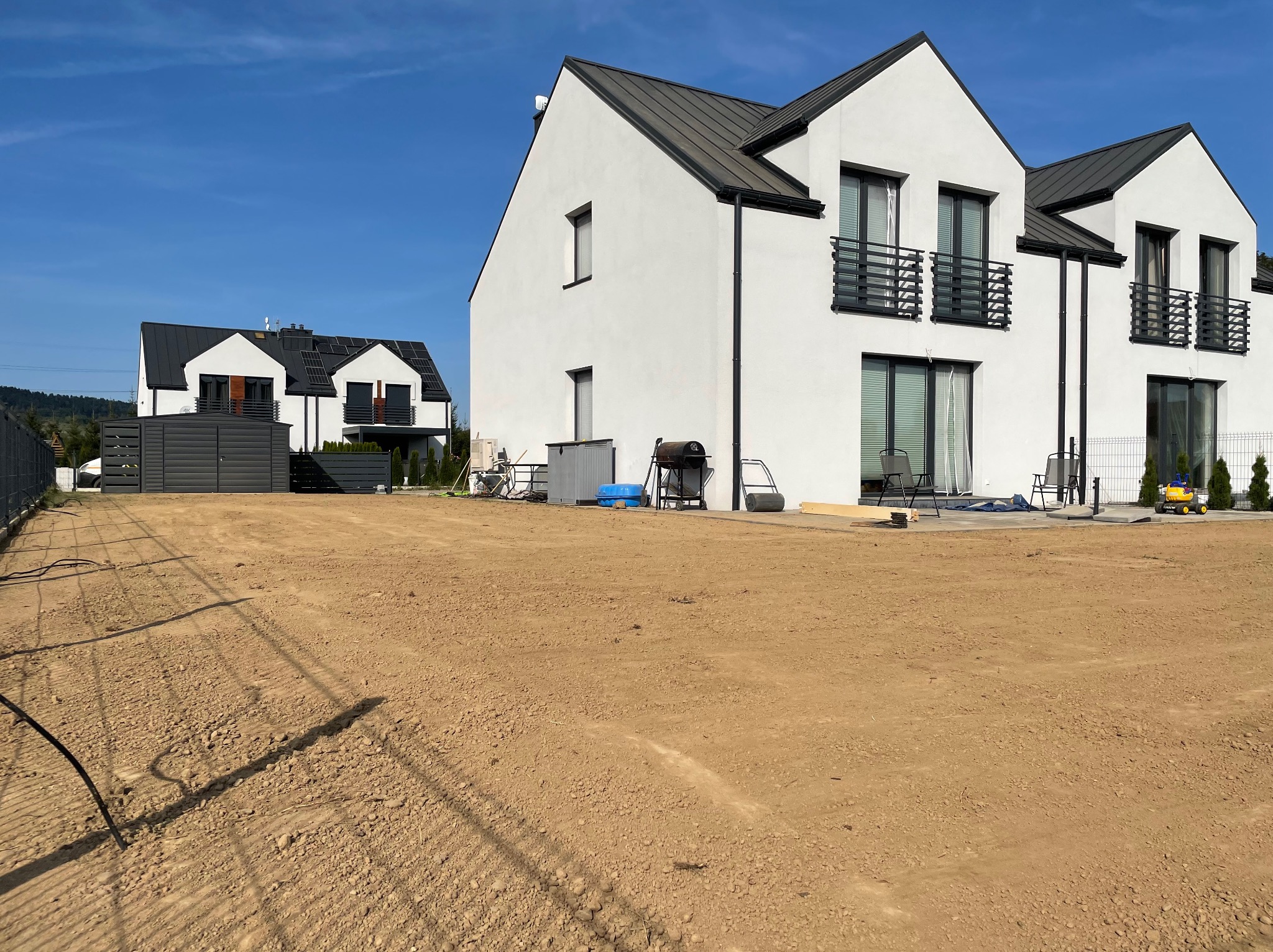 Przygotowany teren pod budowę nowoczesnego domu z widoczną świeżo wyrównaną ziemią, sprzętem budowlanym i sąsiednimi domami w tle, na tle błękitnego nieba.