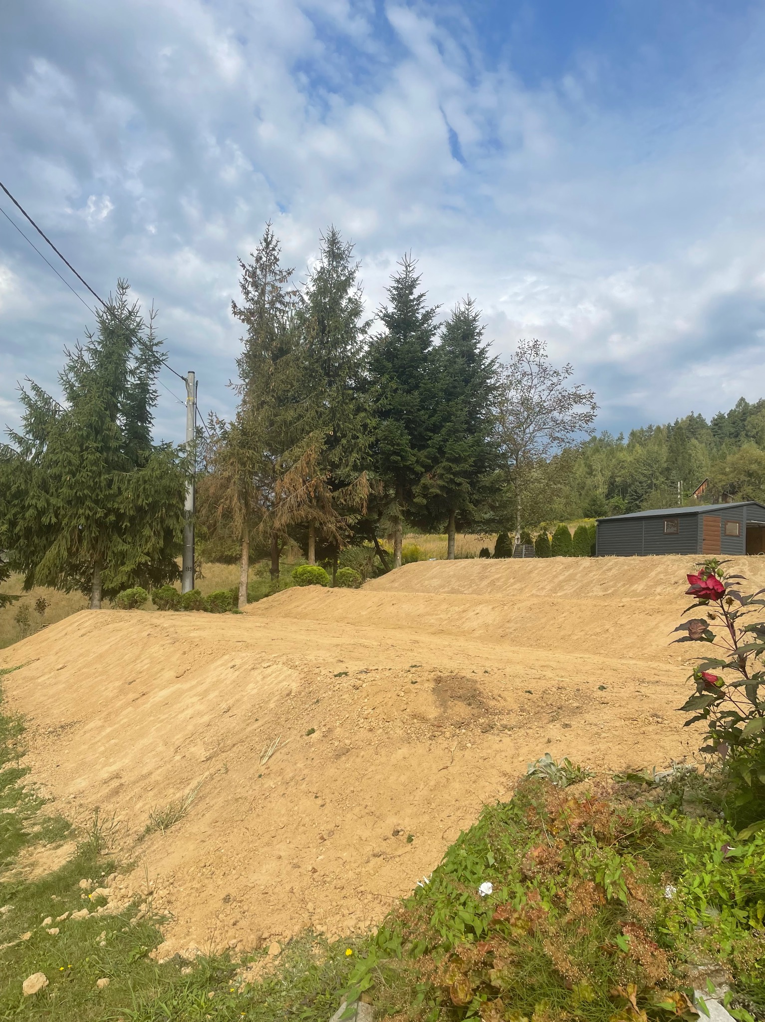 Ukształtowany teren z górkami i dolinami, przygotowany pod budowę lub dalsze prace ziemne, z widocznymi drzewami i budynkiem gospodarczym w tle, częściowo zachwaszczony.