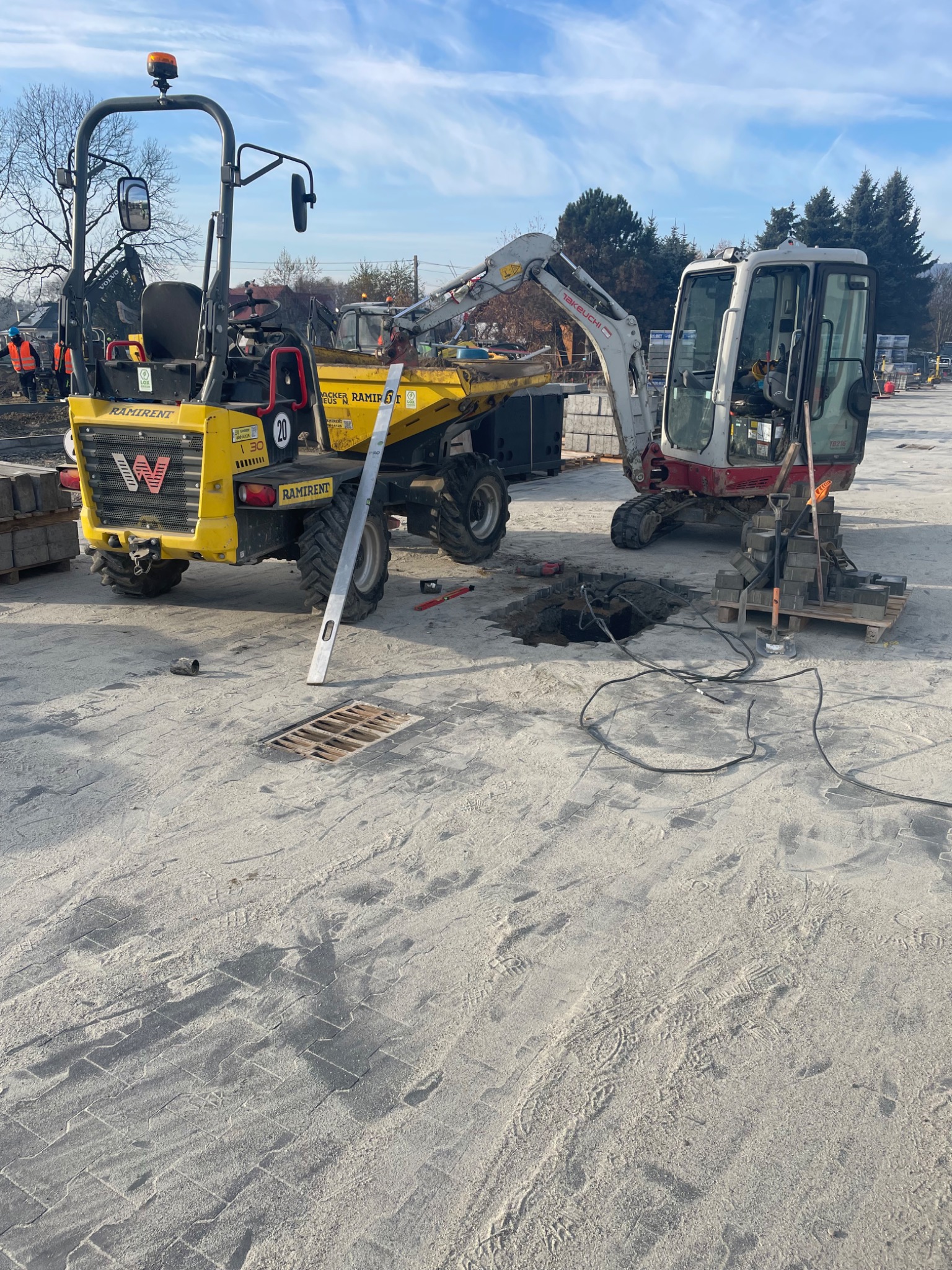 Żółty wozidło i mini koparka Takeuchi TB216 na placu budowy z kostką brukową, widoczny wykop z kablami i studzienka kanalizacyjna, poziomica oparta o koło wozidła.