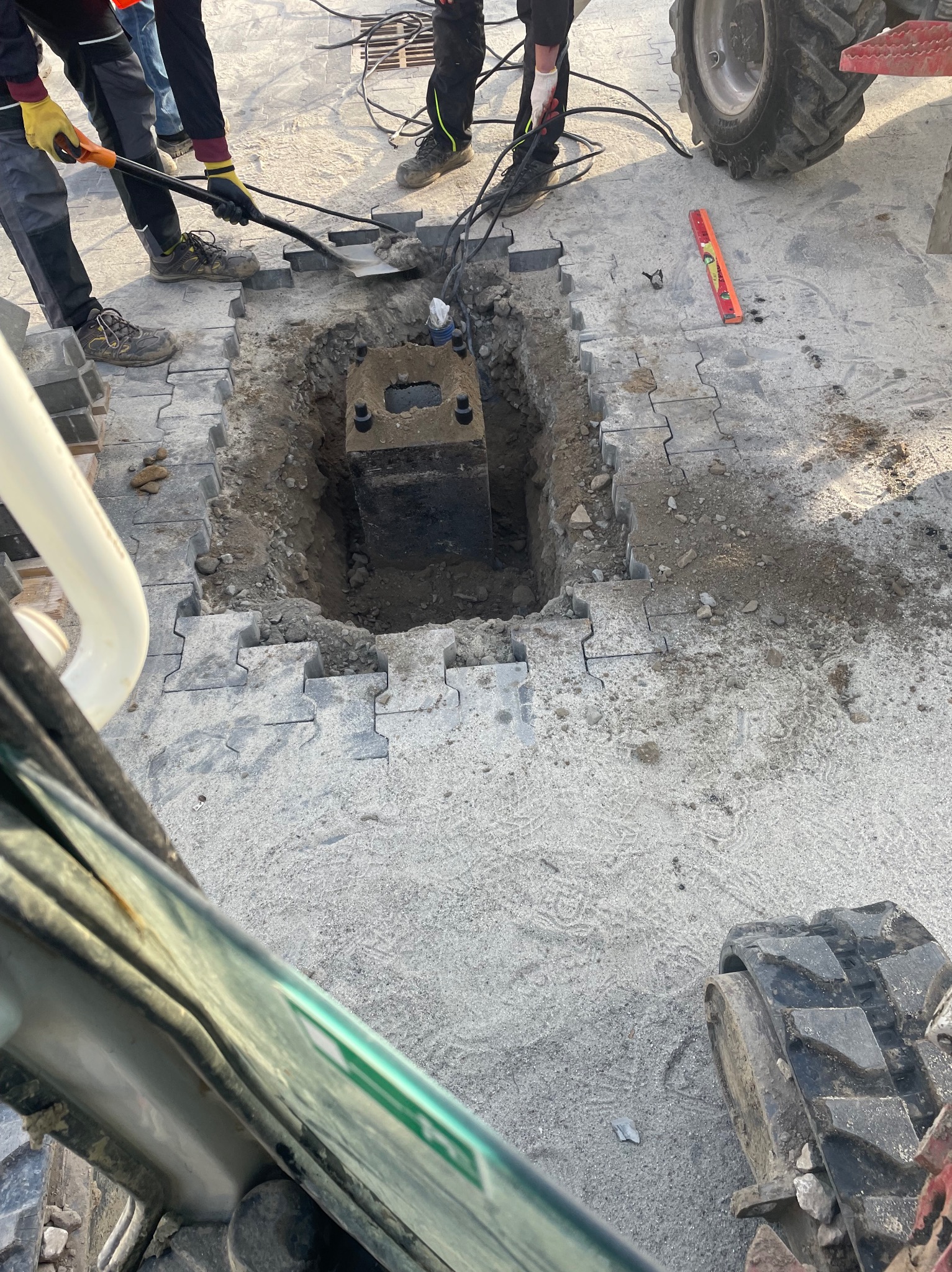 Ekipa budowlana kopie dół pod betonowy element na placu wyłożonym szarą kostką, widoczny fragment traktora i poziomica.
