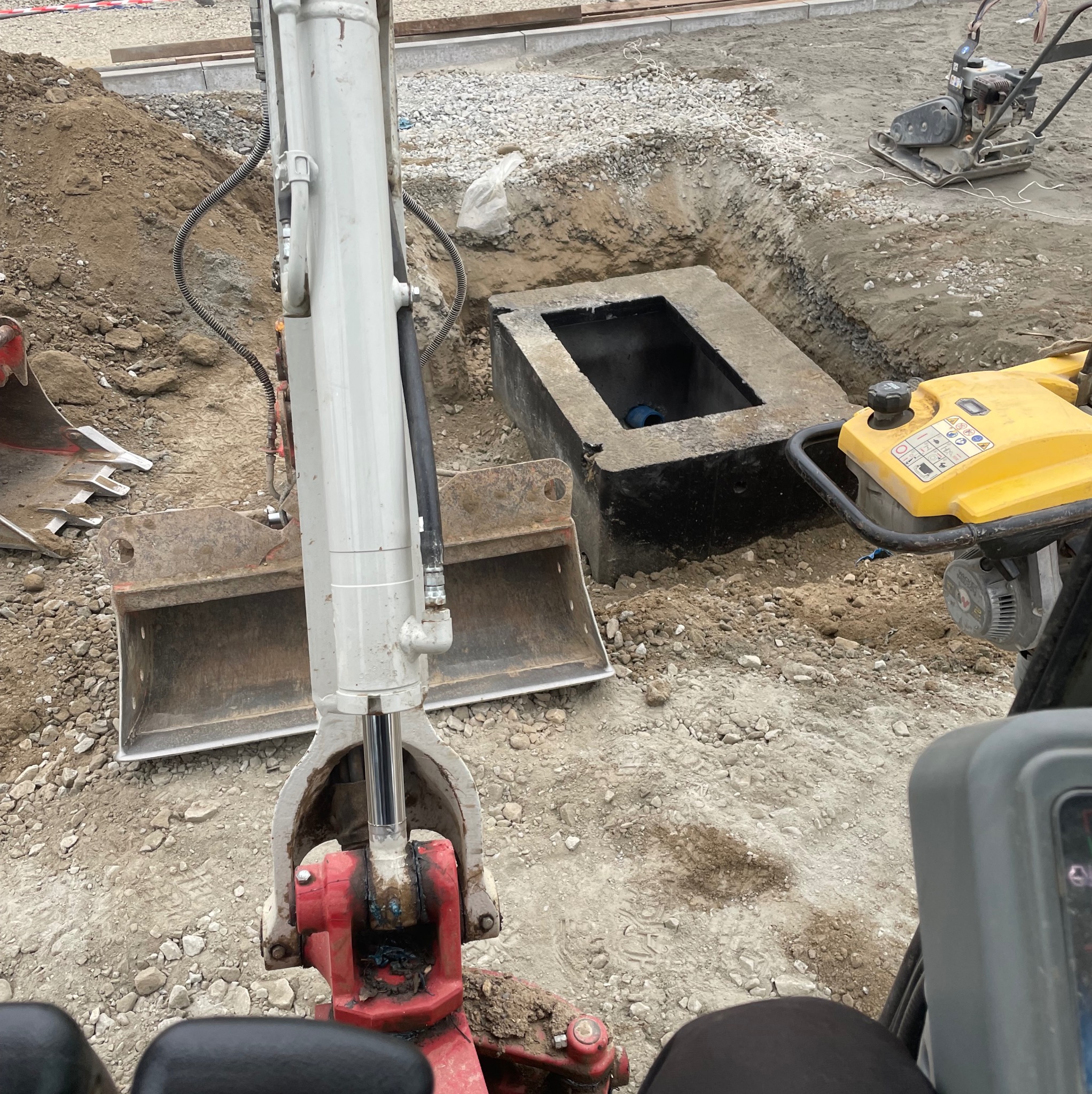 Wykopany dół z betonową studzienką, widok z kabiny minikoparki, ubijarka wibracyjna w tle.