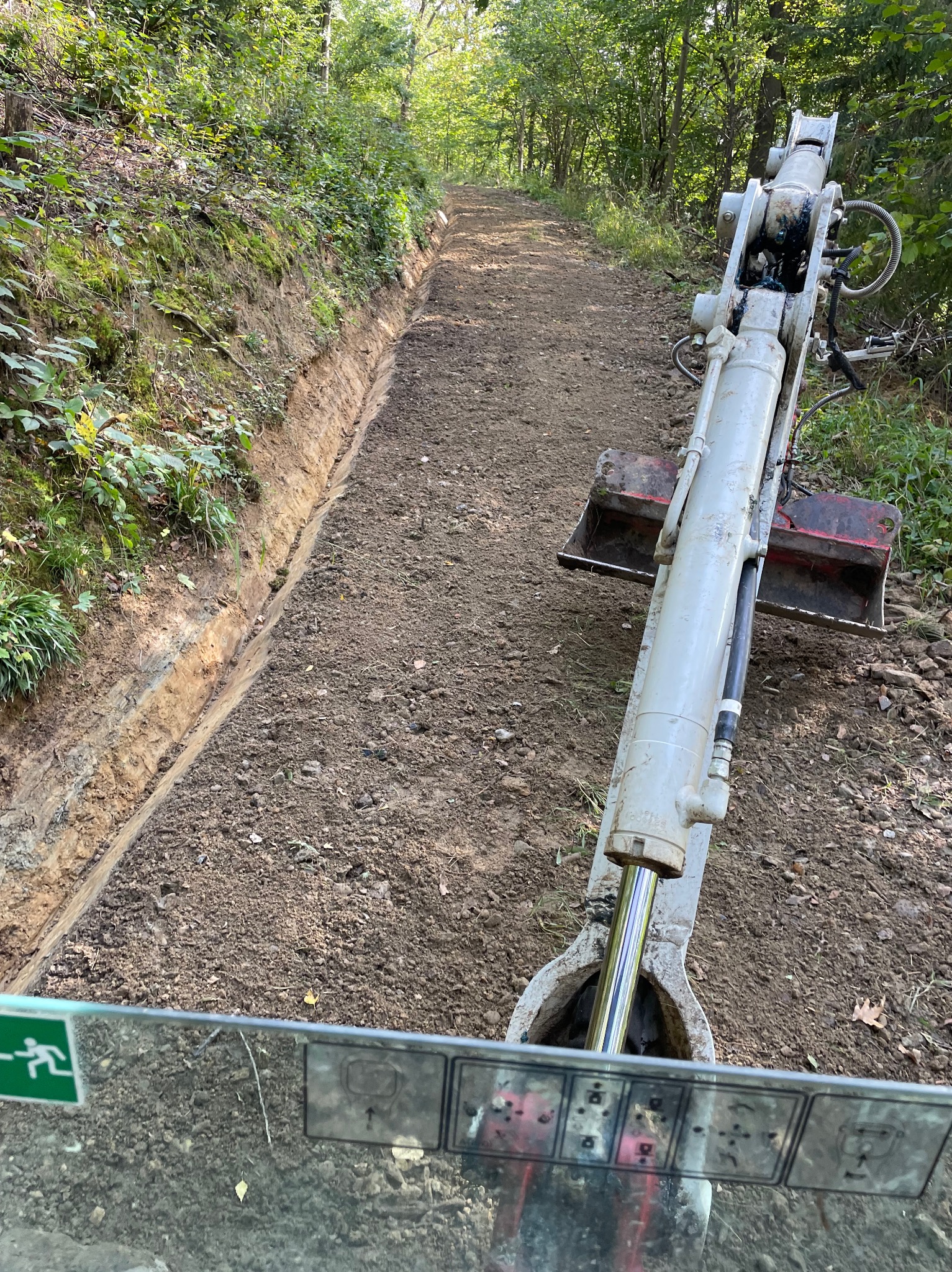 Wąski wykop w zalesionym terenie, widok z kabiny koparki na ramię z łyżką i przygotowany teren pod drogę.