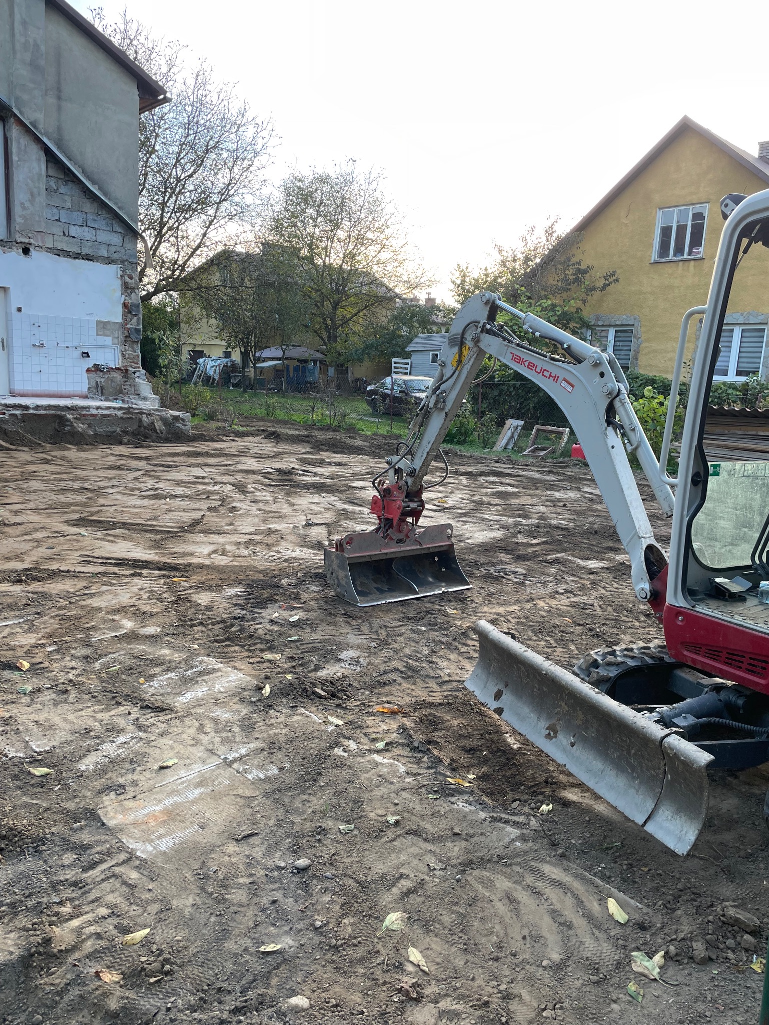 Mała koparka Takeuchi z łyżką i lemieszem na placu budowy, widoczne ślady opon w błotnistym gruncie, w tle częściowo zburzony budynek i dom jednorodzinny.