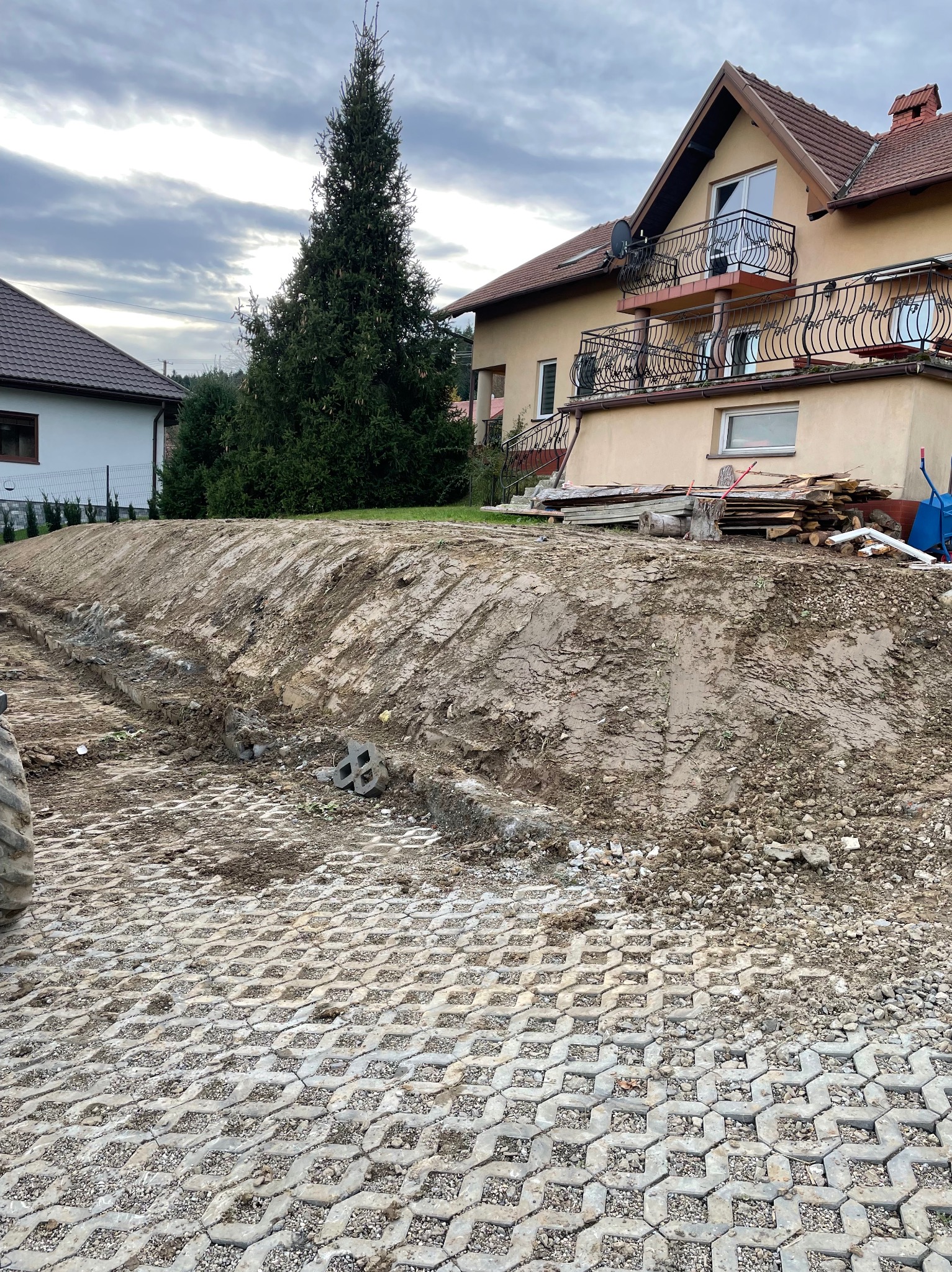 Przygotowanie terenu pod budowę domu: wyrównany teren z widocznymi śladami opon, ułożona częściowo ażurowa kostka brukowa, w tle dom z balkonem i sterta drewna.