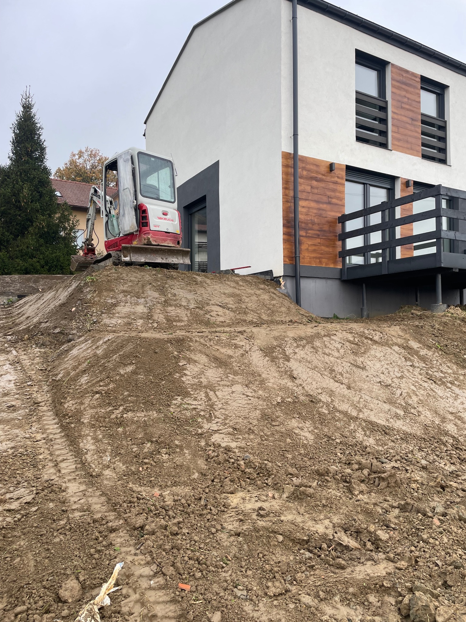 Mała koparka Takeuchi na nierównym, gliniastym terenie przed nowoczesnym domem z drewnianą elewacją i czarnymi ramami okiennymi w pochmurny dzień.