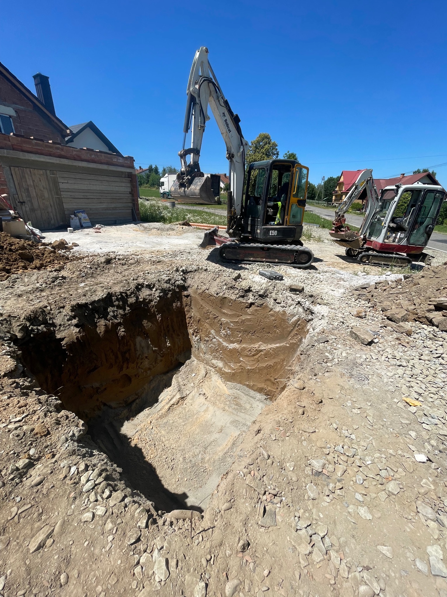 Głęboki wykop w ziemi, obok dwie koparki Bobcat, w tle budynek i błękitne niebo. Prace ziemne w toku, widoczny przekrój wykopu z warstwami gleby.