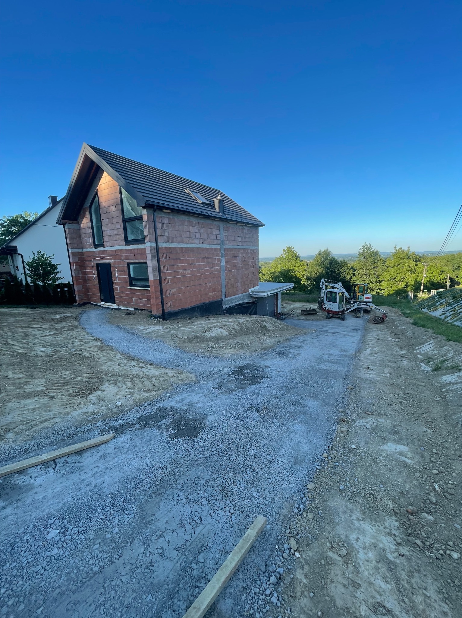 Budowa domu w Chełmcu: stan surowy otwarty, widoczne dwie koparki i utwardzona droga dojazdowa. Dom z cegły z czarną stolarką okienną na tle błękitnego nieba.