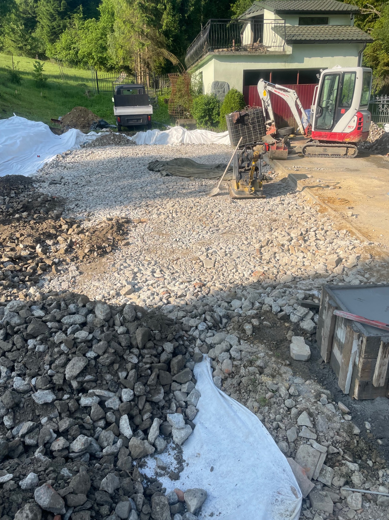 Etap utwardzania terenu pod budowę: ubity tłuczeń, mini koparka, ubijarka wibracyjna, przygotowane formy betonowe. Biała plandeka zabezpiecza teren.