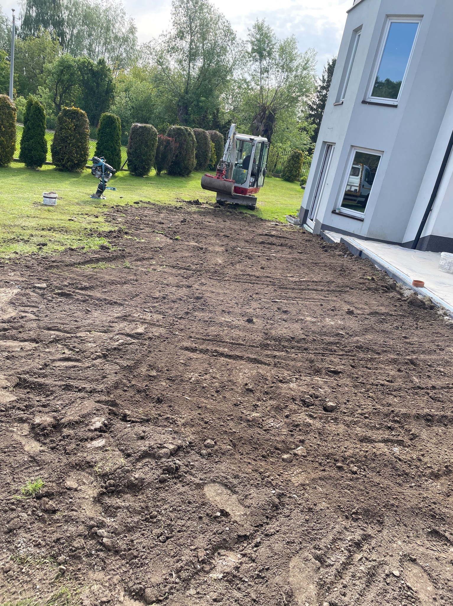 Przygotowany teren pod budowę obok domu jednorodzinnego. Widoczny mini-koparka i ubijak wibracyjny. Ślady opon na ziemi. Częściowo położony chodnik.