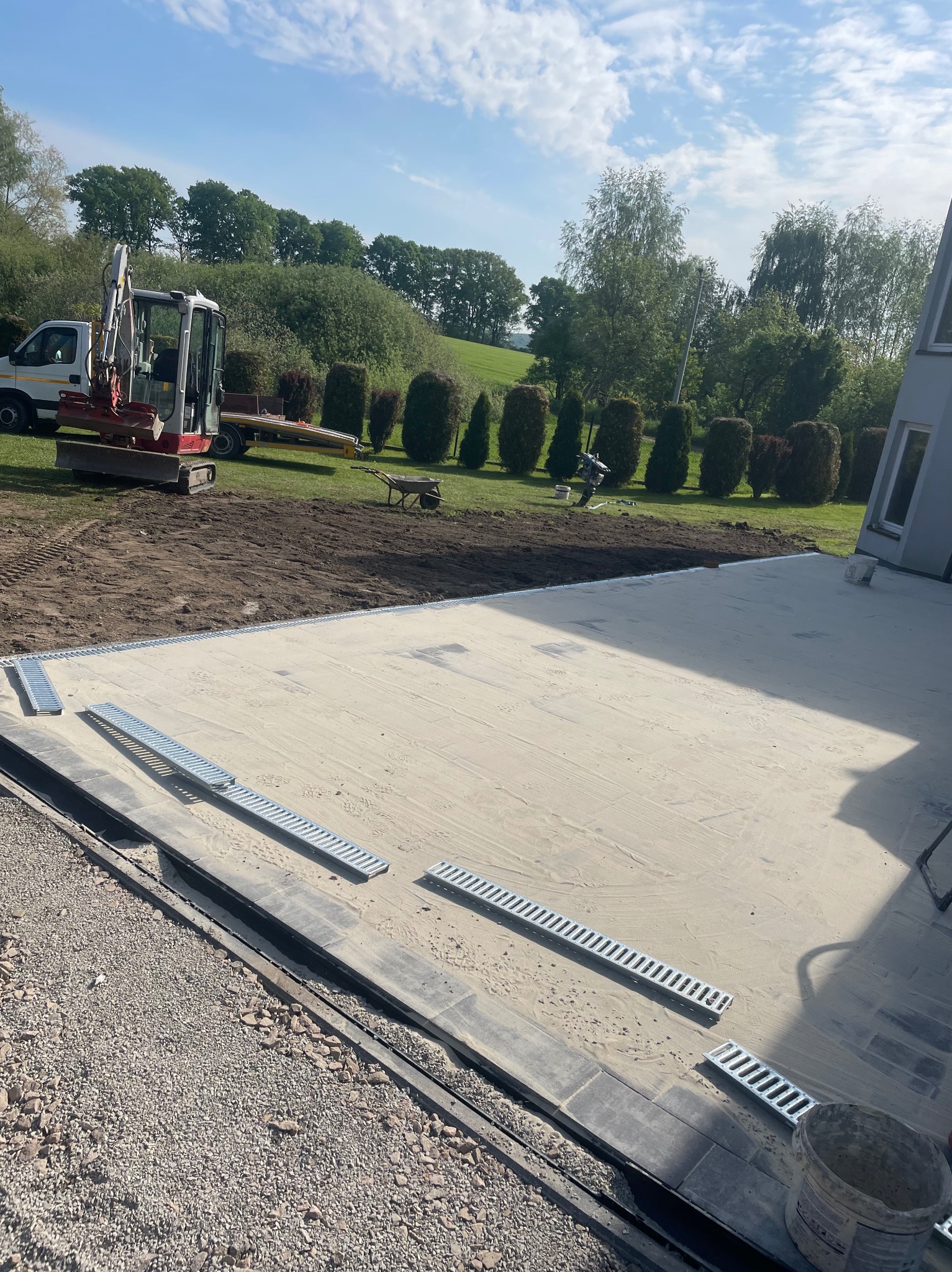 Przygotowany teren pod wylewkę z widocznym drenażem liniowym; w tle minikoparka i uporządkowany teren zielony. Jasne, słoneczne oświetlenie.