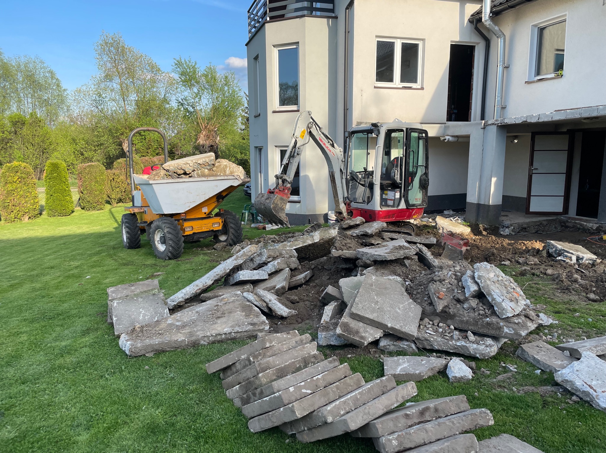 Mini koparka i wywrotka na trawniku obok budynku w trakcie prac ziemnych. Stos gruzu i płyt betonowych. Widok na dom z oknami i drzewa w tle. Słoneczny dzień.