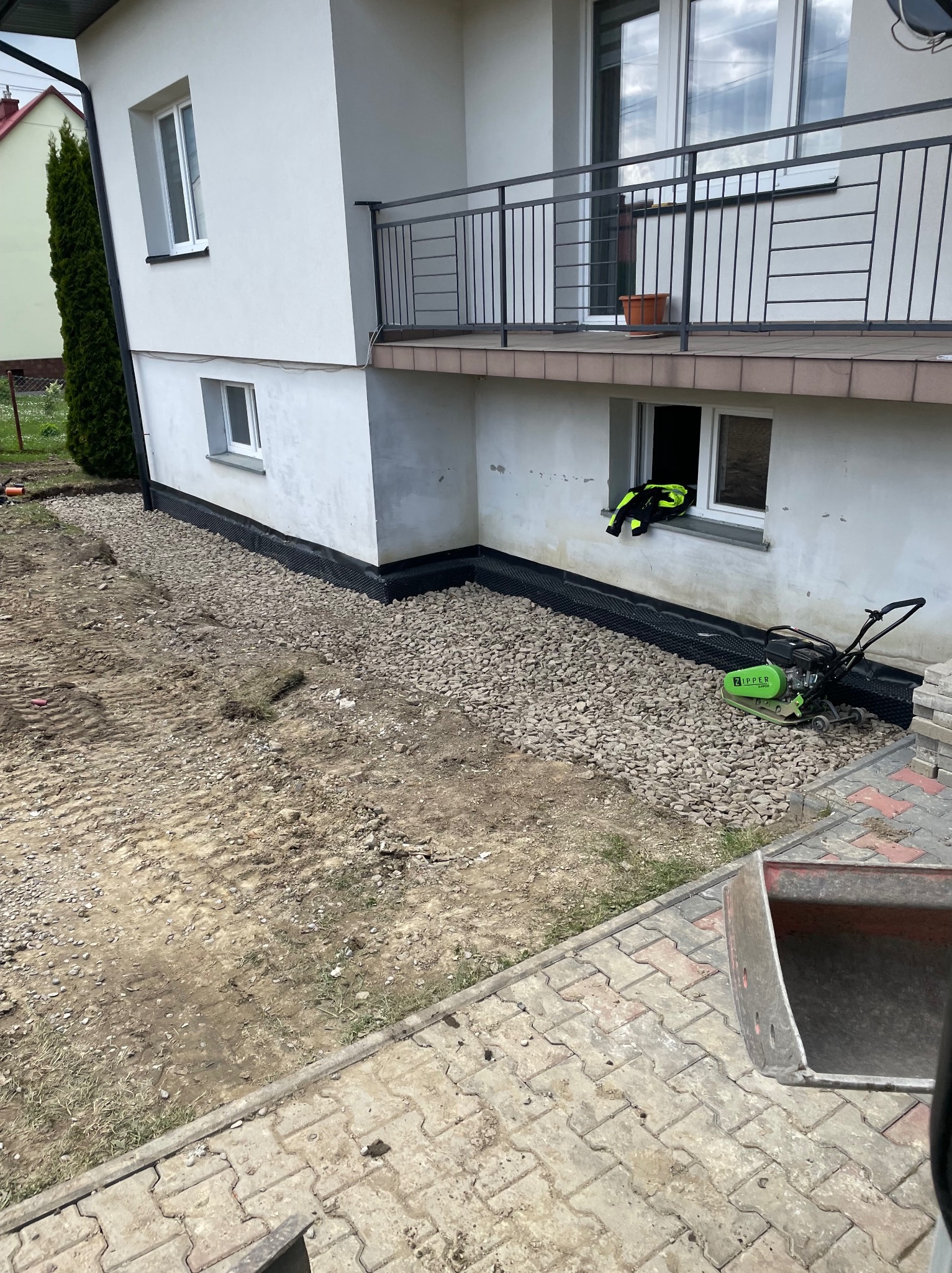 Czarna folia izolacyjna przy fundamencie domu, obsypana żwirem. Obok stoi zagęszczarka. Widoczny fragment chodnika z kostki i łyżka koparki.