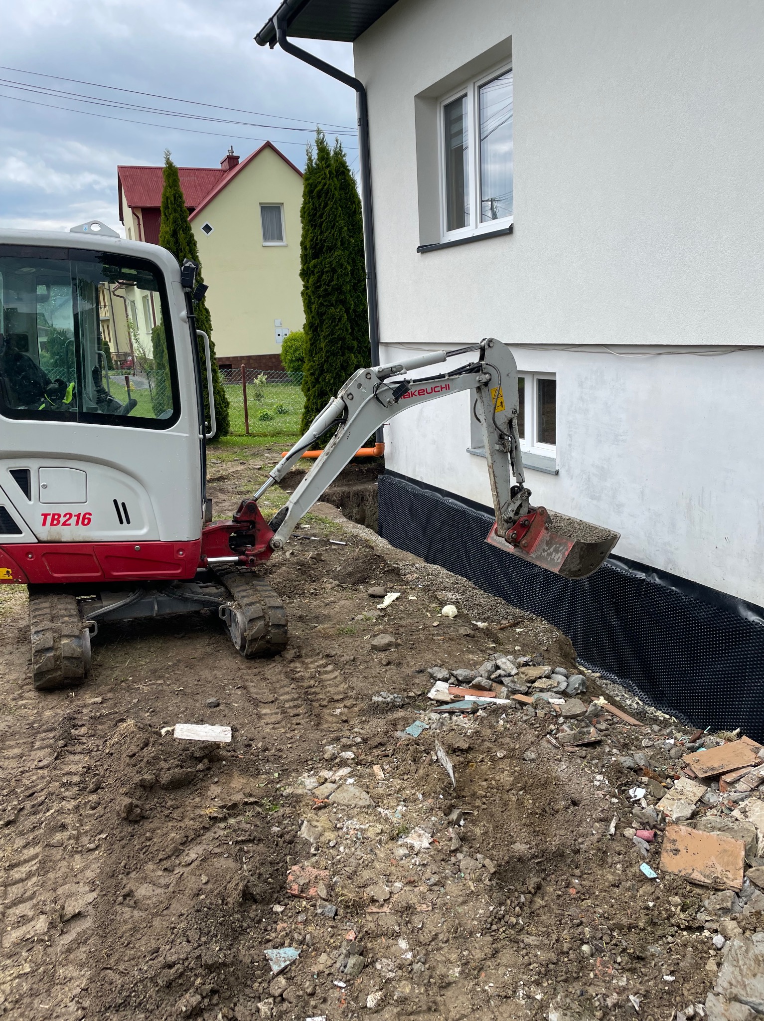 Mini koparka przy wykopie obok domu z izolacją fundamentów. Widoczne ślady opon w błocie i gruz. Prace ziemne w toku, przygotowanie pod drenaż.