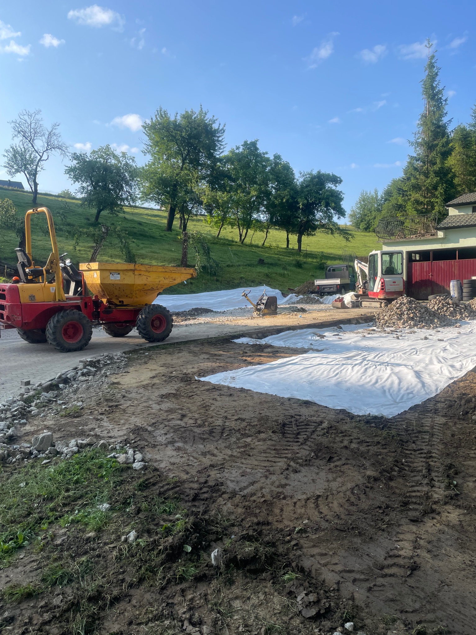 Żółto-czerwona wywrotka na placu budowy z białym materiałem i kamieniami, w tle zieleń wzgórz i błękitne niebo. Etap prac przygotowawczych.