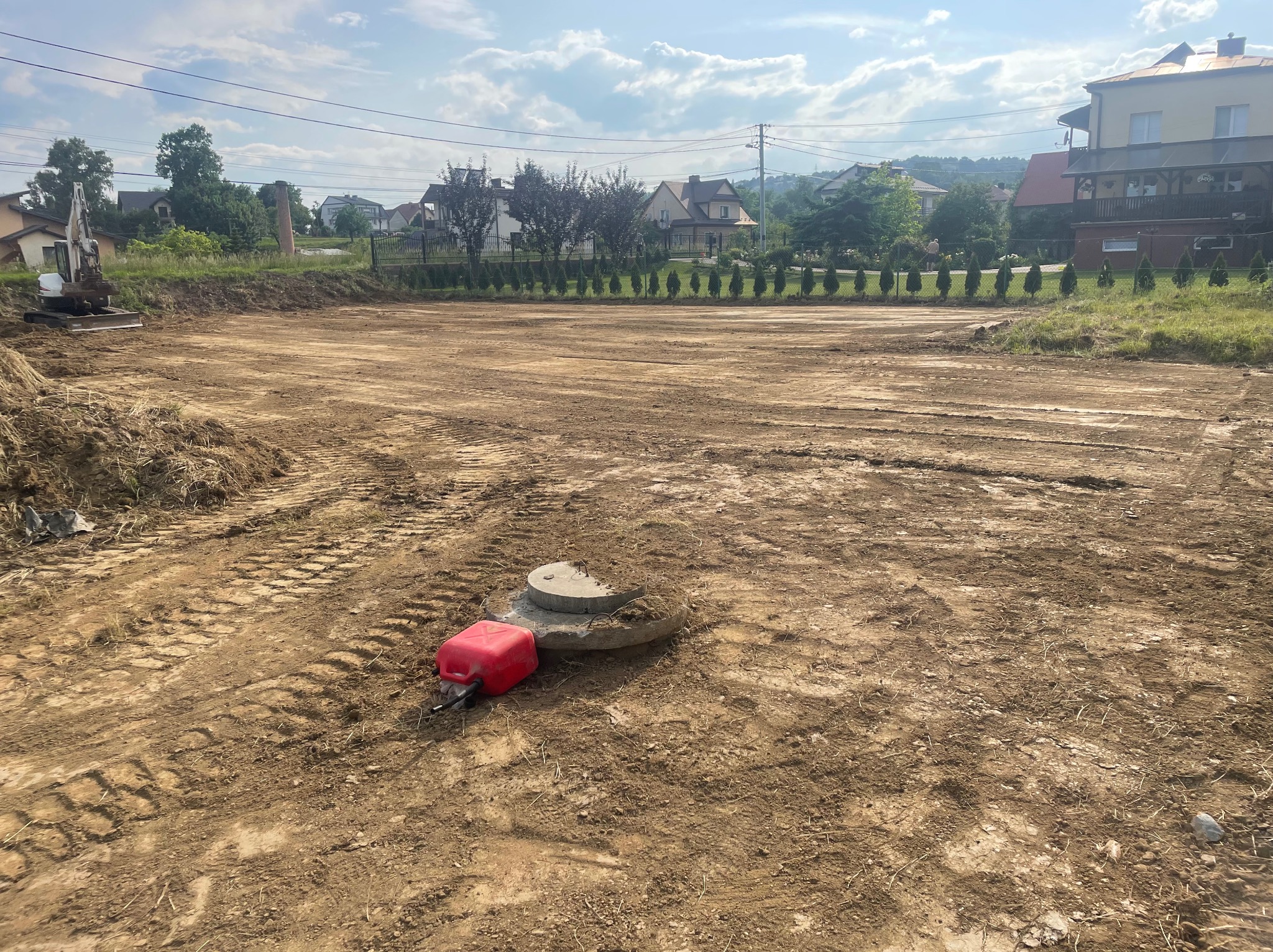 Przygotowany teren pod budowę z widocznymi śladami opon i małą koparką w tle, w słoneczny dzień. Widoczny czerwony kanister i betonowy element.