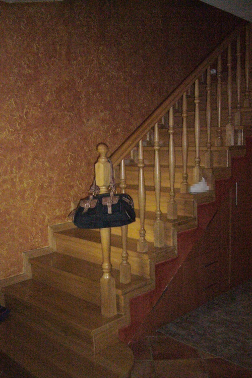 Drewniane schody wewnętrzne z toczonymi balustradami i brązową torbą zawieszoną na słupku, ściana z teksturą w odcieniach brązu.