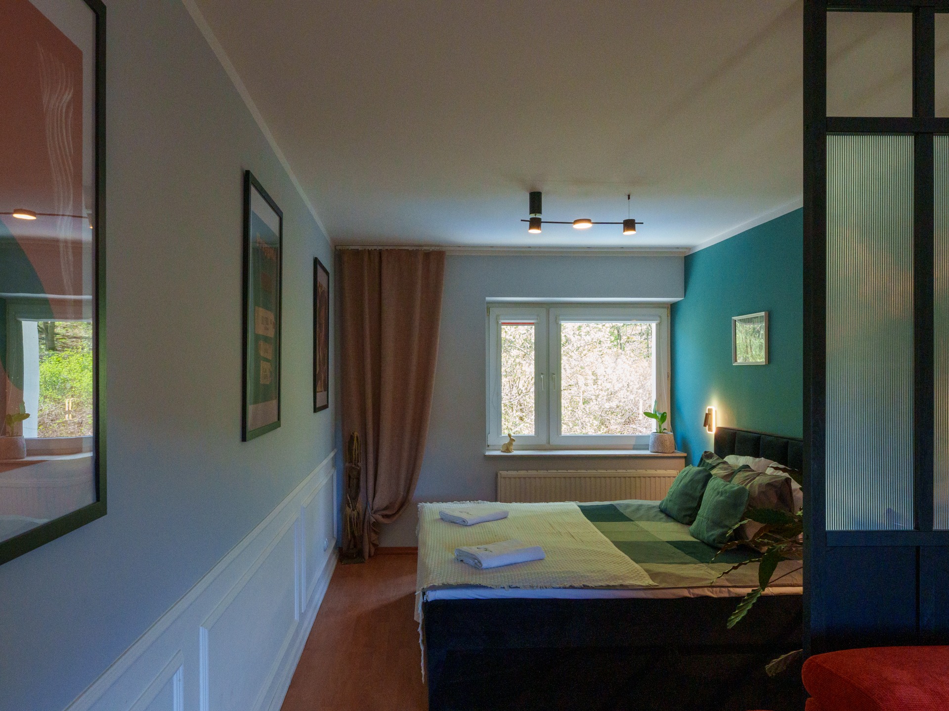 Przytulna sypialnia z łóżkiem, zielonymi poduszkami i pledem w paski, jasnymi ścianami z dekoracyjną sztukaterią i oknem z widokiem na zieleń.