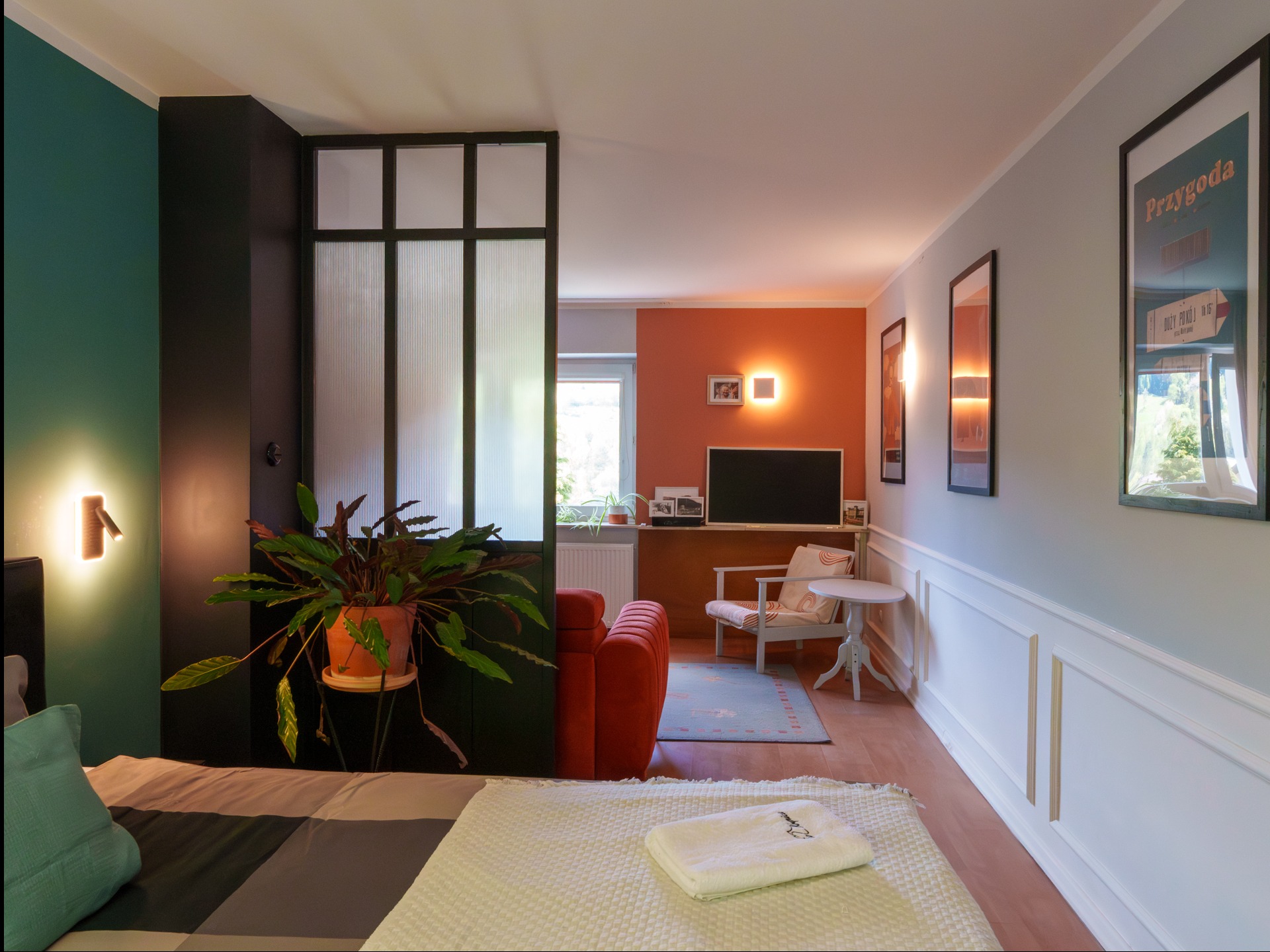 Nowoczesne wnętrze mieszkania z podziałem na strefy: sypialnia z rośliną, salon z sofą i TV. Aranżacja z dbałością o detale, ciepłe oświetlenie, plakaty na ścianie.