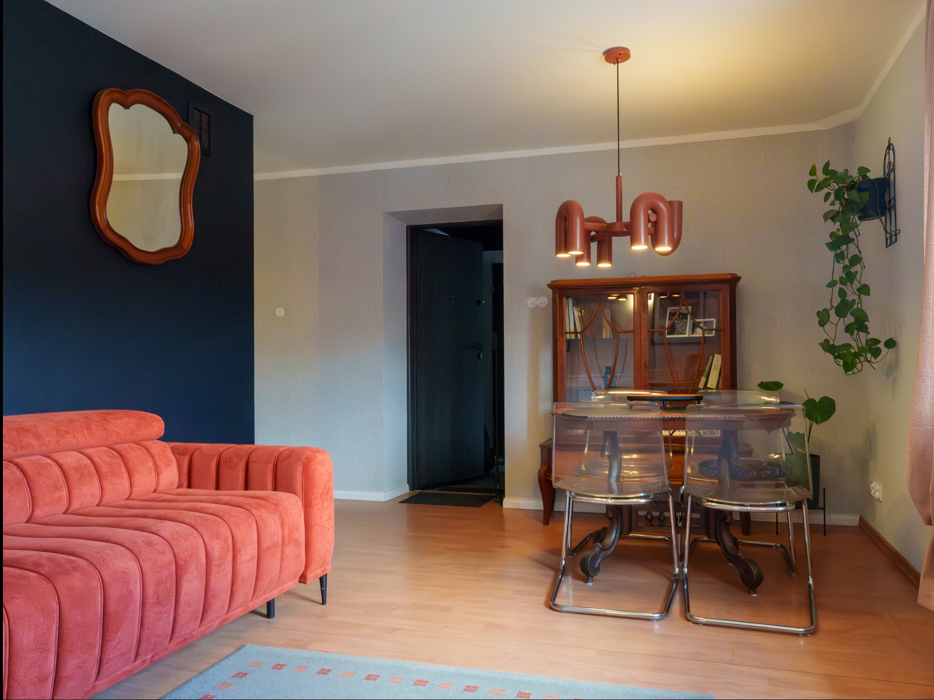 Salon z koralową sofą na tle granatowej ściany, drewniane meble i lampa w stylu retro. Lustro o nieregularnym kształcie dodaje unikalnego charakteru wnętrzu.