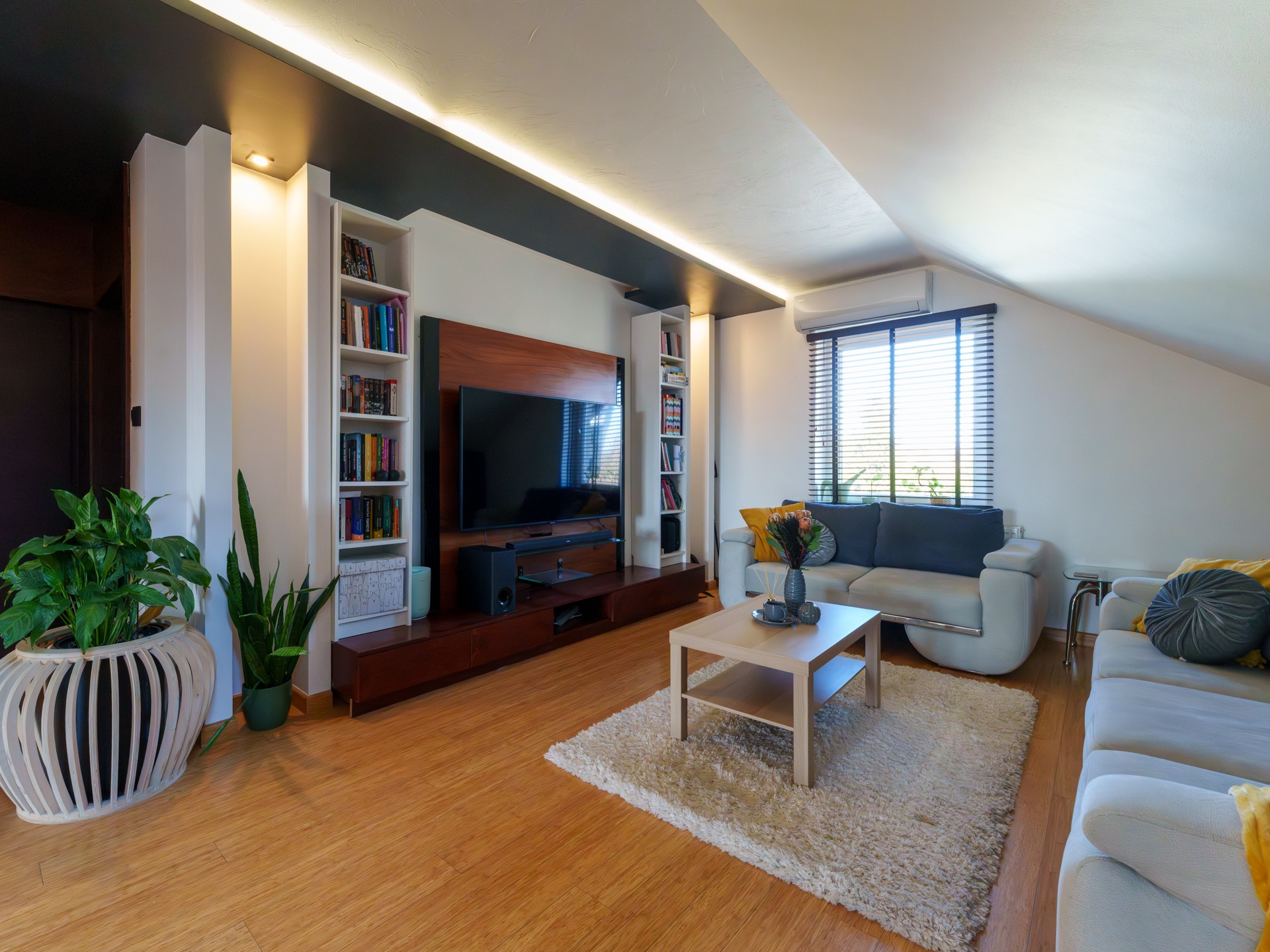 Nowoczesny salon na poddaszu z drewnianą podłogą, białymi regałami i telewizorem, doświetlony listwami LED. Widok z dołu na jasne wnętrze.