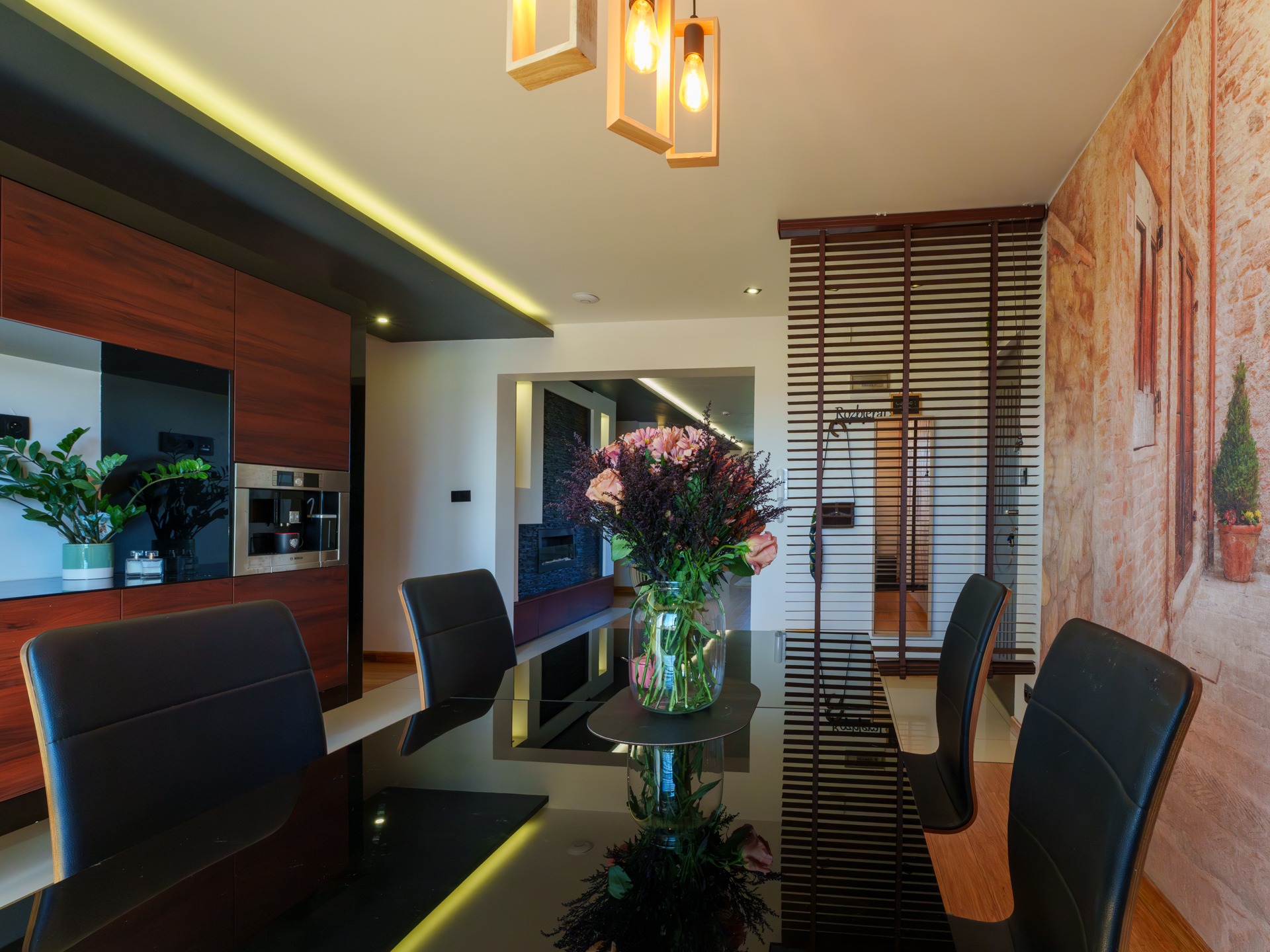 Nowoczesna jadalnia: czarny stół z bukietem róż, skórzane krzesła, zabudowa kuchenna z ekspresem, designerskie lampy i fototapeta z motywem toskańskim.