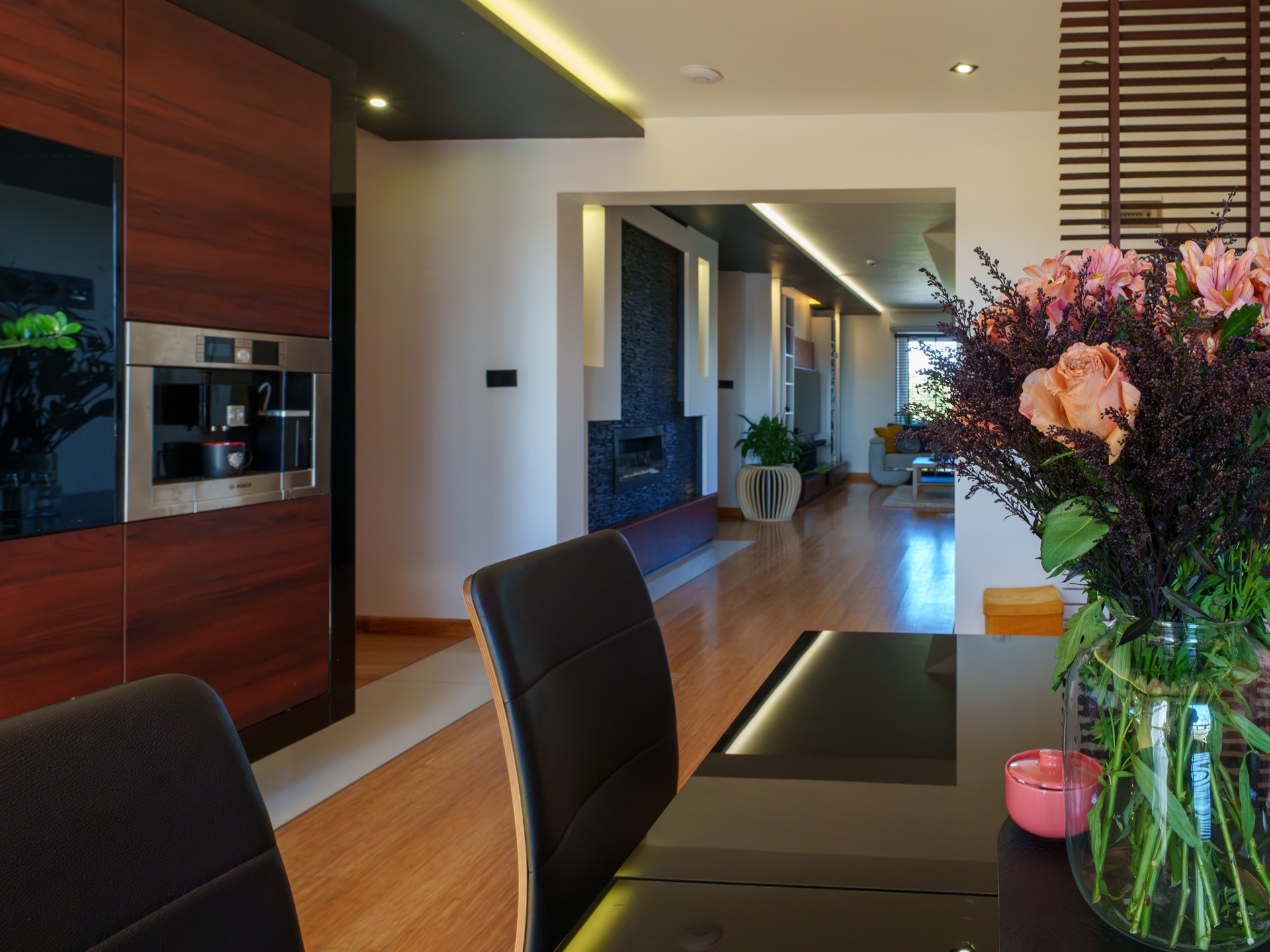 Nowoczesne wnętrze: parkiet, kwiaty w wazonie na stole, zabudowa kuchenna z ekspresem do kawy, kominek z czarnym kamieniem, oświetlenie ledowe.