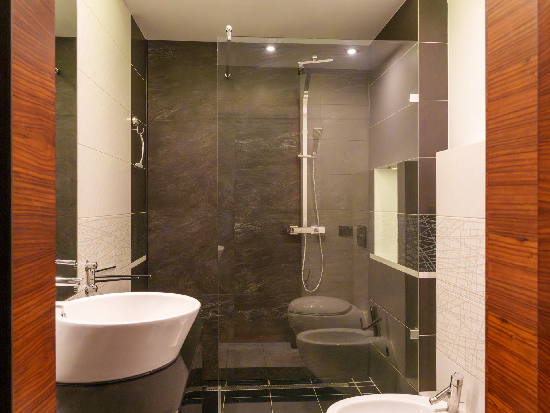 Elegancka łazienka z kabiną prysznicową typu walk-in, umywalką nablatową i bidetem. Ciemne płytki marmurowe kontrastują z białymi elementami i drewnianym wykończeniem.