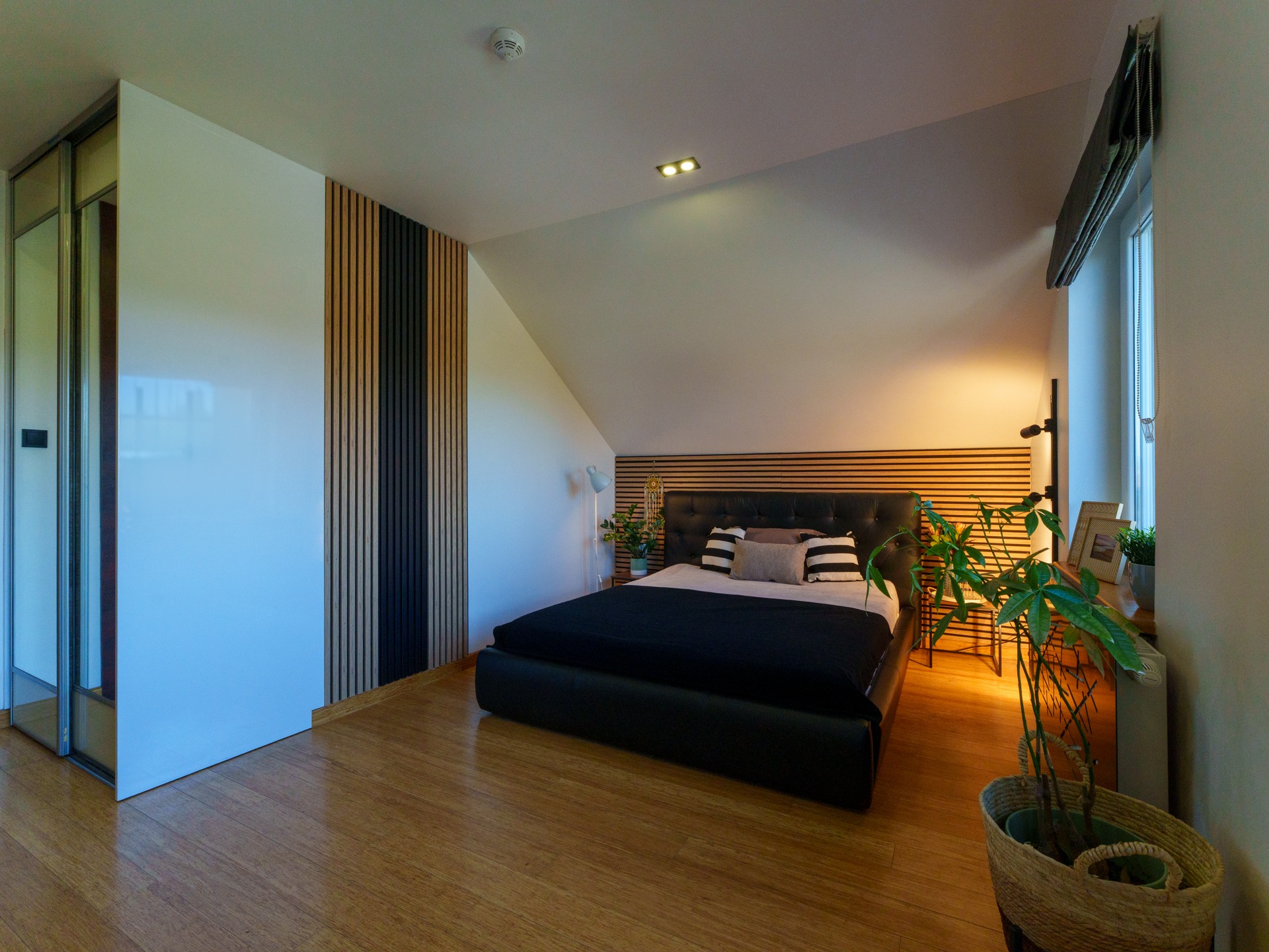 Jasna sypialnia na poddaszu z drewnianą podłogą i panelami ściennymi. Czarne łóżko z pikowanym zagłówkiem, roślina doniczkowa i okno z roletą rzymską.