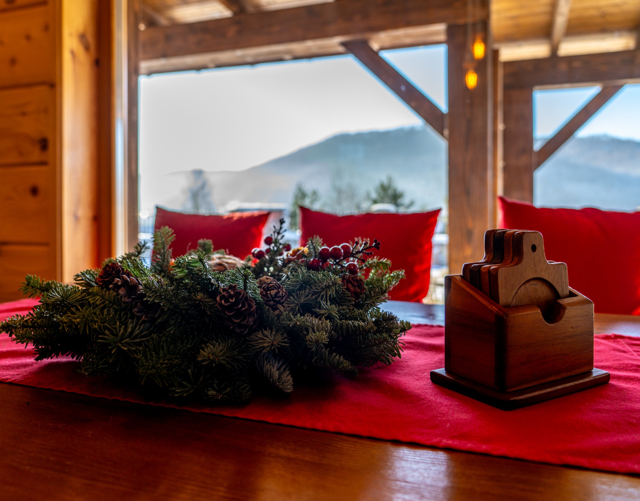 Dekoracja świąteczna z gałązek świerku i szyszek na czerwonym bieżniku, drewniane podkładki pod napoje w tle okna z widokiem na góry.