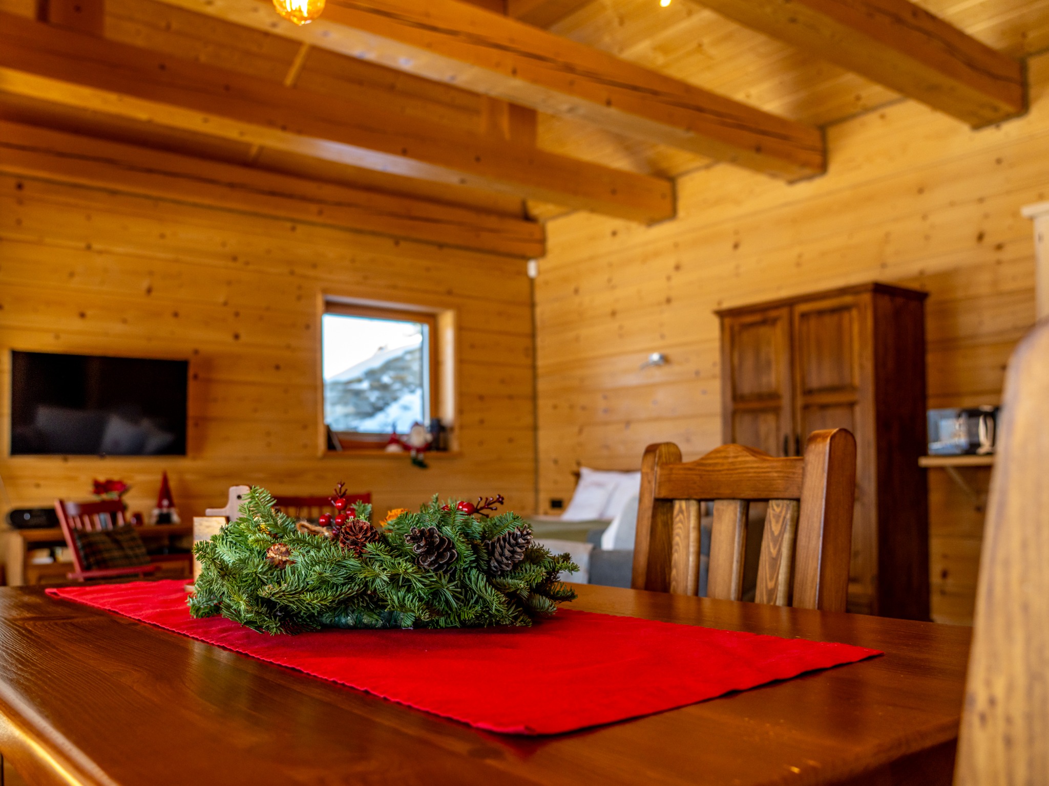 Rustykalne wnętrze z drewnianymi ścianami i sufitem. Na stole dekoracyjny wieniec z igliwia i szyszek na czerwonym bieżniku. W tle okno z zimowym krajobrazem.